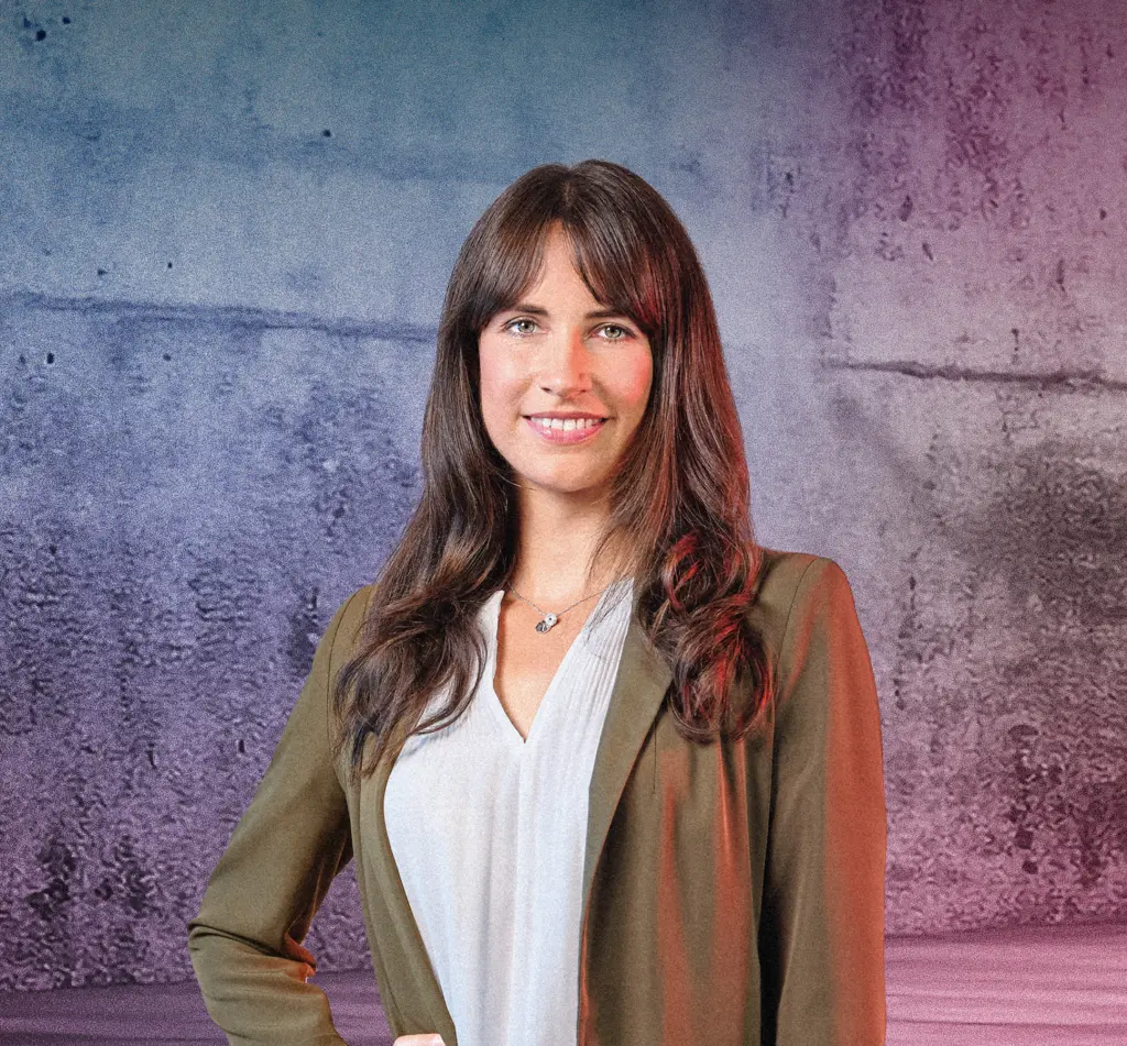 Eine Person steht lächelnd vor einer Betonwand mit blau-pinkem Lichtverlauf. Rechts im Bild leuchten mehrere diagonale Neonlichtstreifen. Die Szene wirkt modern und freundlich.