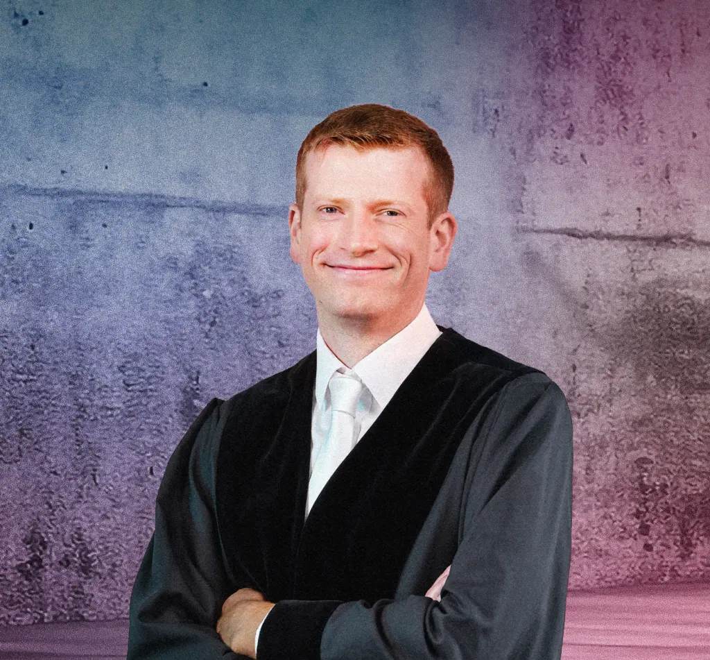 Eine Person in einer Robe steht lächelnd vor einer Betonwand mit farbigem Lichtverlauf in Blau und Pink. Rechts im Bild befinden sich mehrere diagonale Neonlichtstreifen, die in den Raum zeigen. Die Szene wirkt modern und professionell.