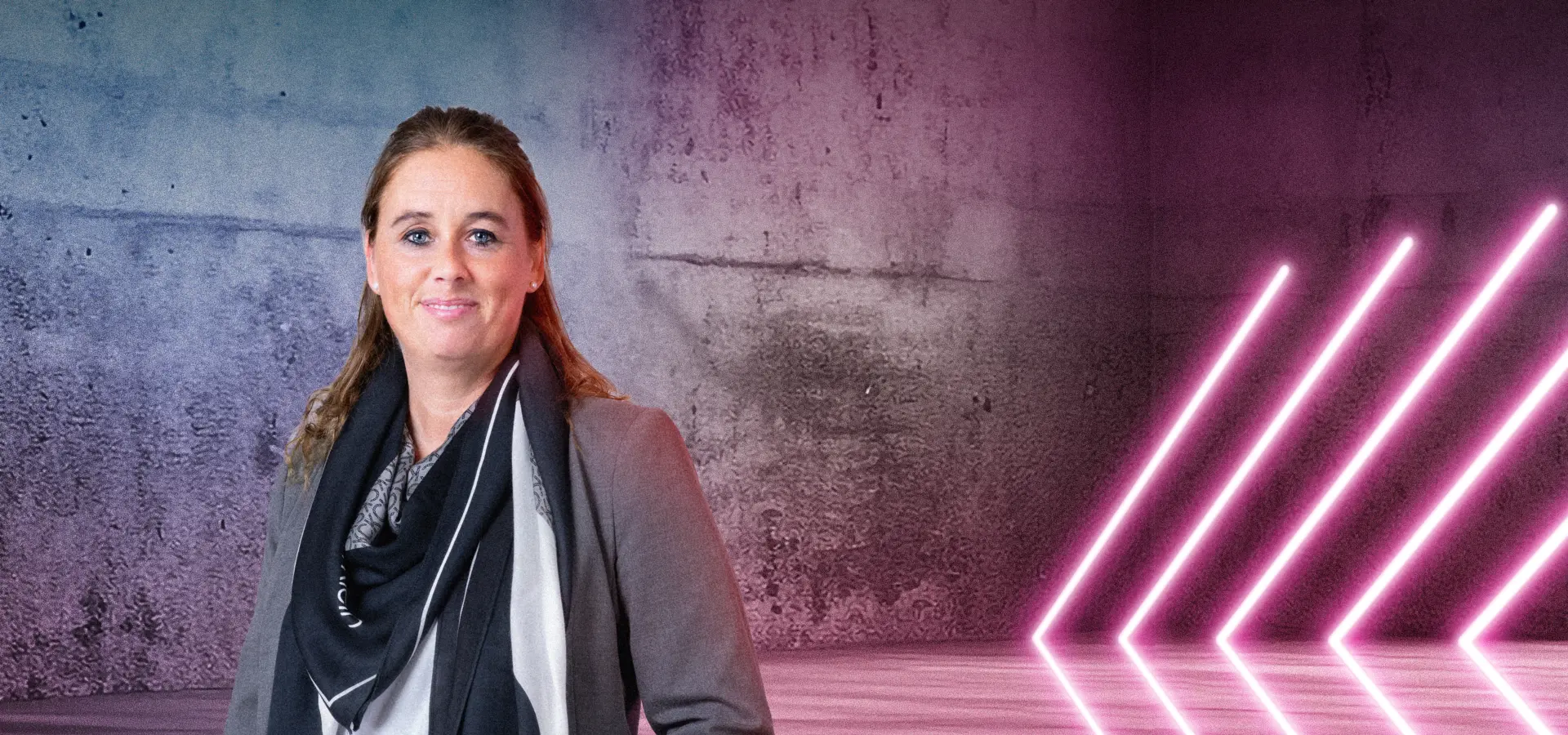 Eine Person steht lächelnd vor einer Betonwand mit blau-pinkem Lichtverlauf. Rechts im Bild leuchten mehrere diagonale Neonlichtstreifen. Die Szene wirkt modern und freundlich.